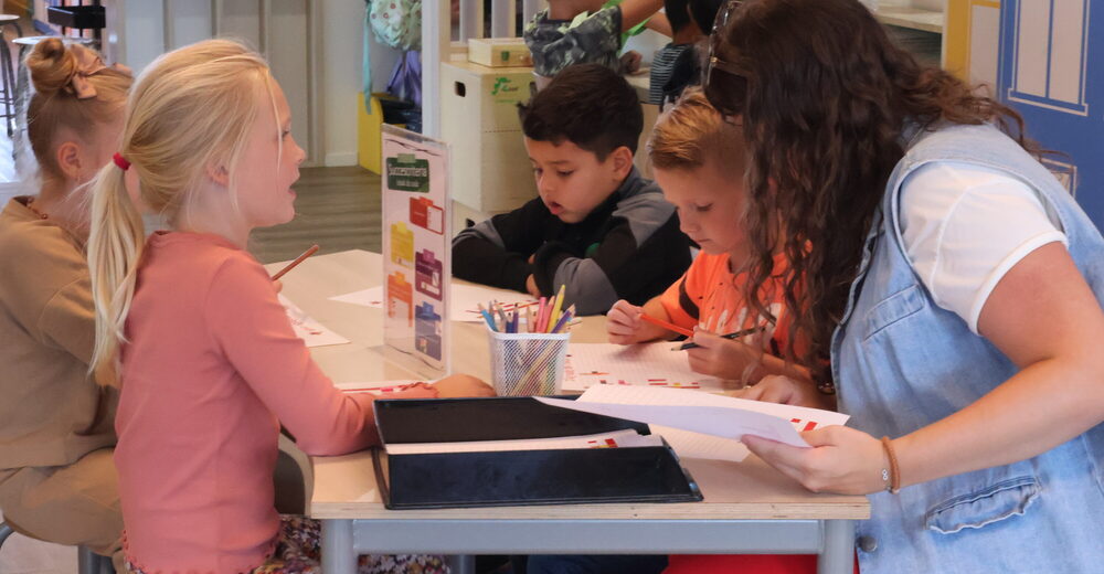 Een juf helpt kinderen aan tafel
